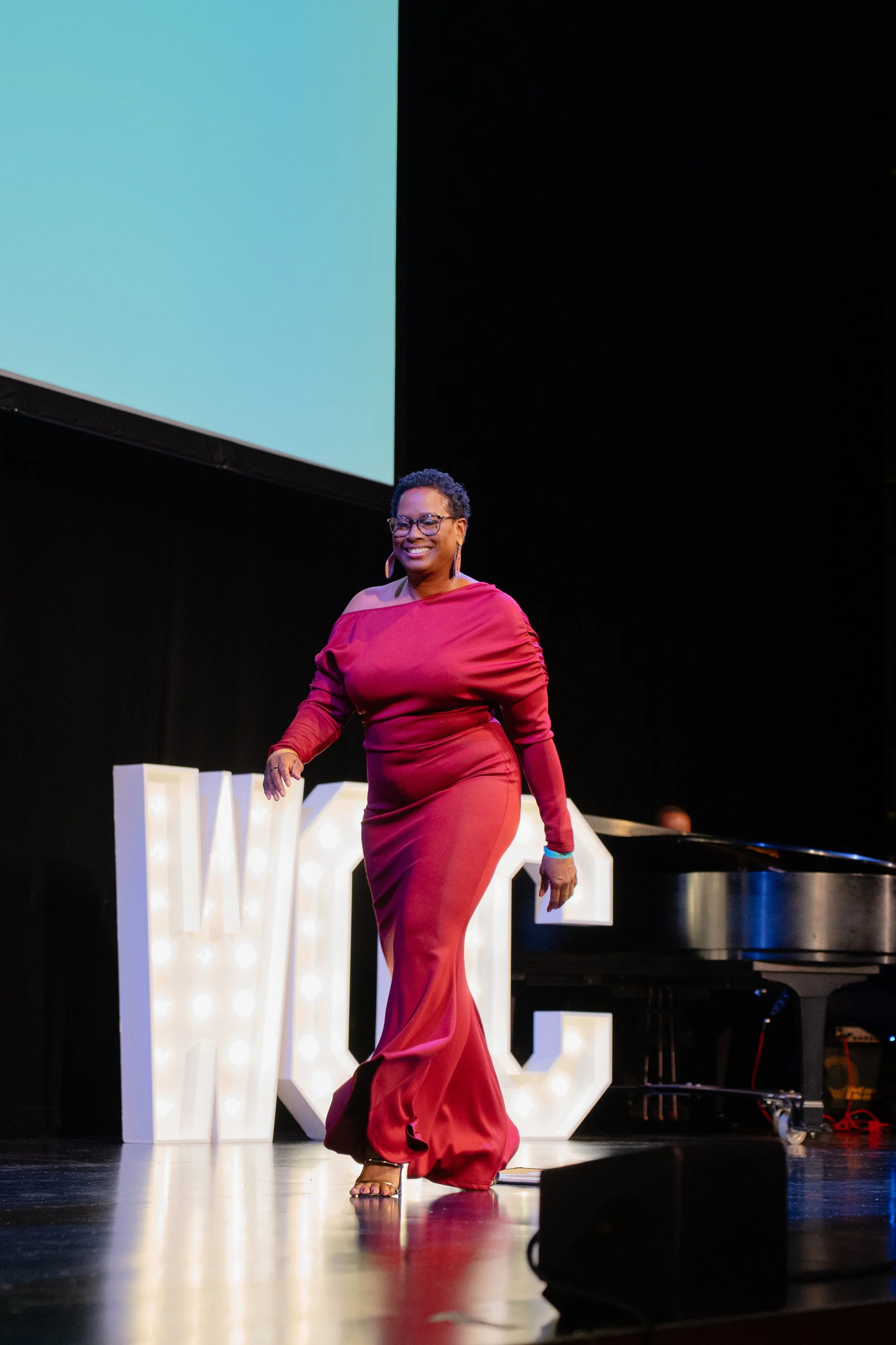 A woman in a red dress with an off-shoulder design walking on stage, smiling, with a large illuminated letter and a piano in the background.