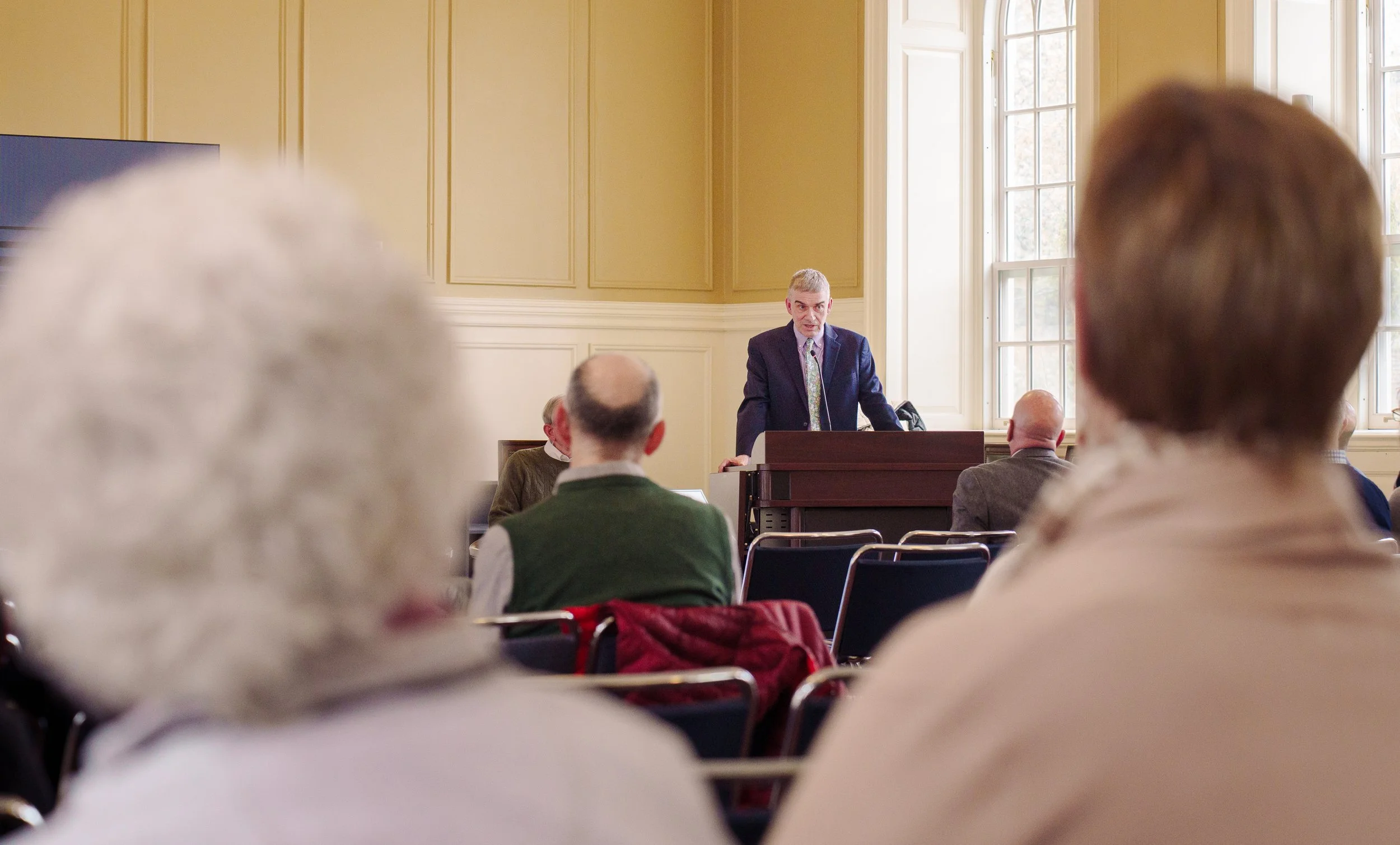 A man in a suit speaking at a podium in a room with large windows and beige walls, while an audience of mostly older adults listens.