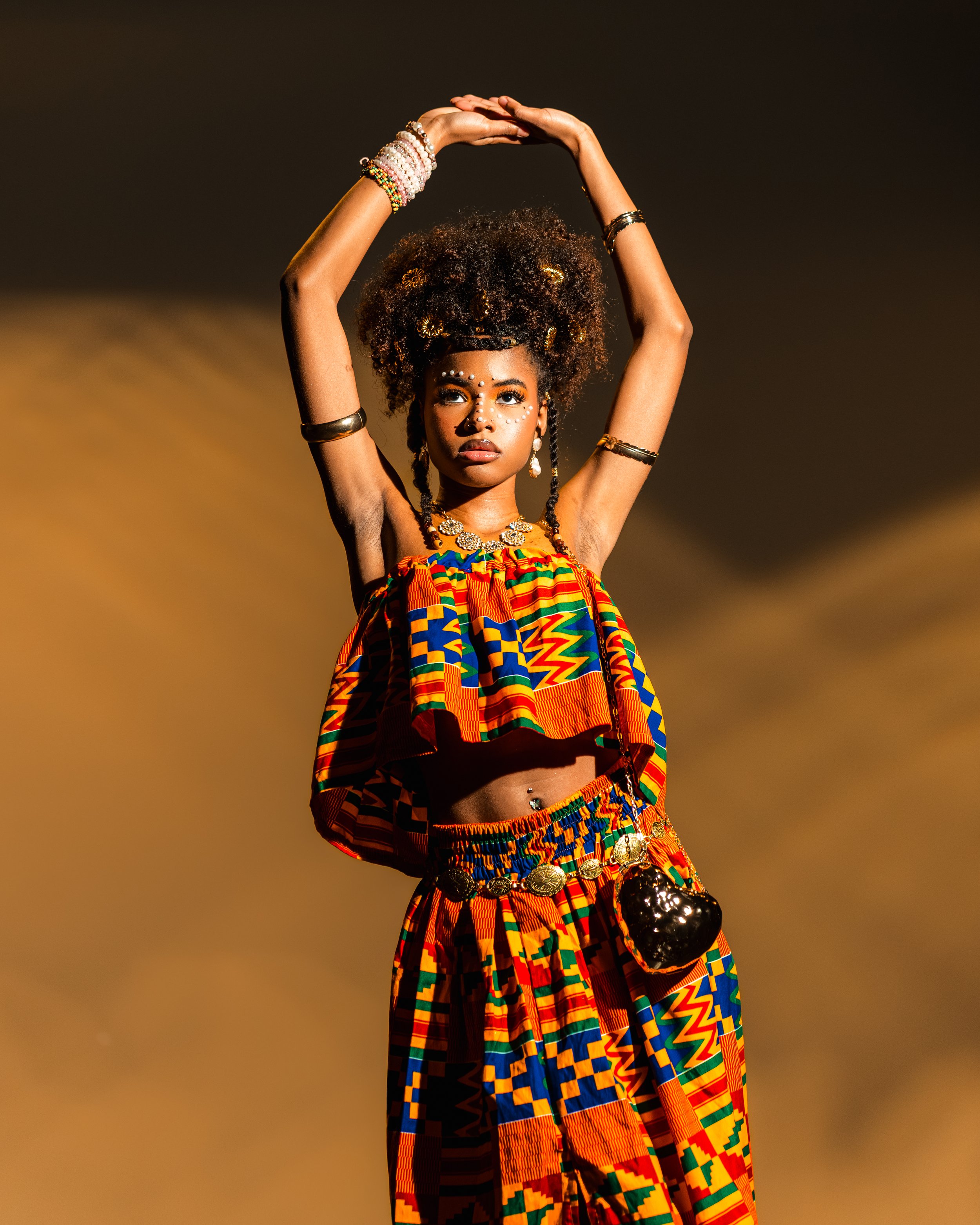 A woman dressed in traditional brightly colored African clothing, accessorized with jewelry and face paint, with her arms raised above her head and long, curly hair.