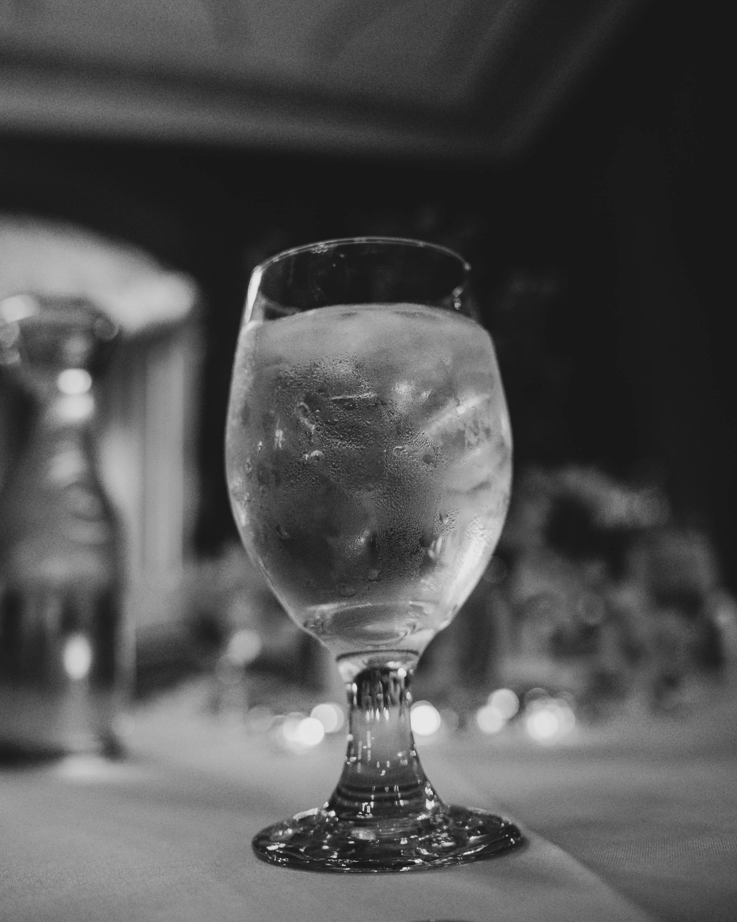 A close-up black and white photograph of a wine glass filled with ice water, with condensation on the outside, set on a table.