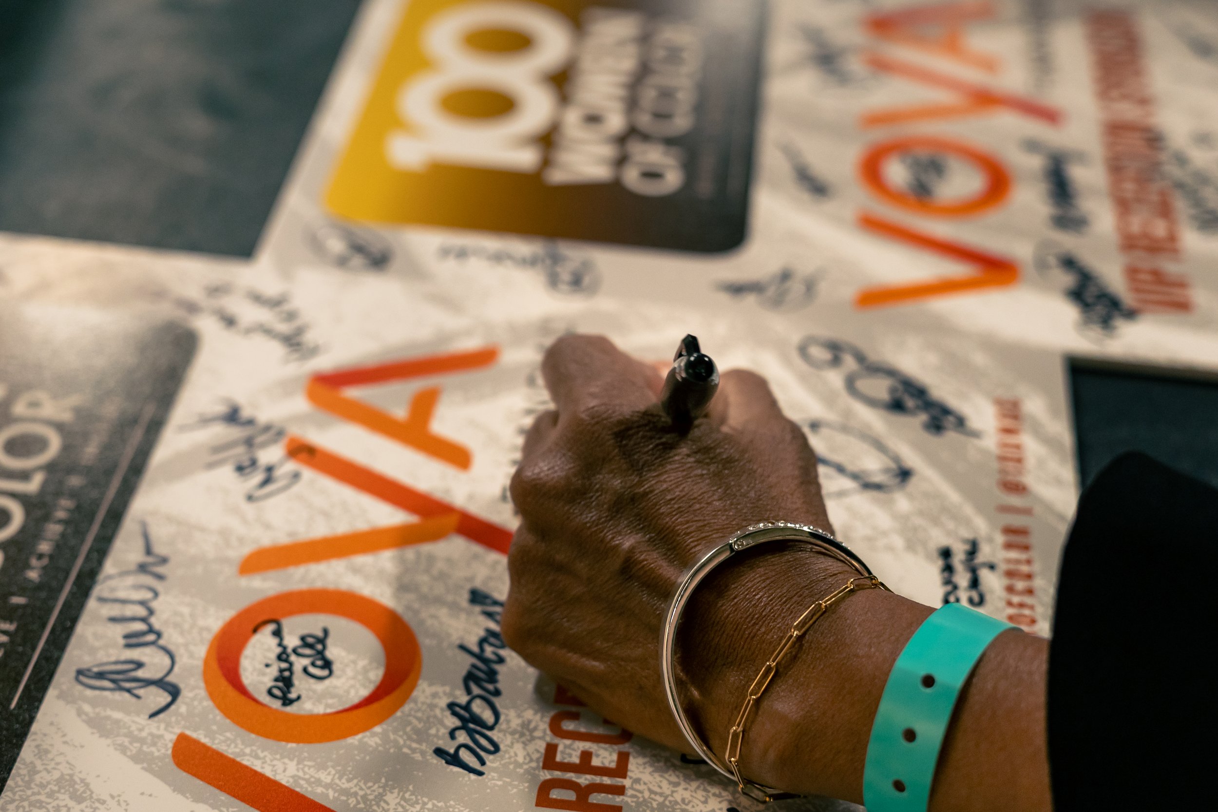 A person's hand signing a poster at what appears to be a convention or event. The poster has colorful text and graphics, with the person's wrist adorned with a black bracelet, a gold bracelet, and a blue wristband.