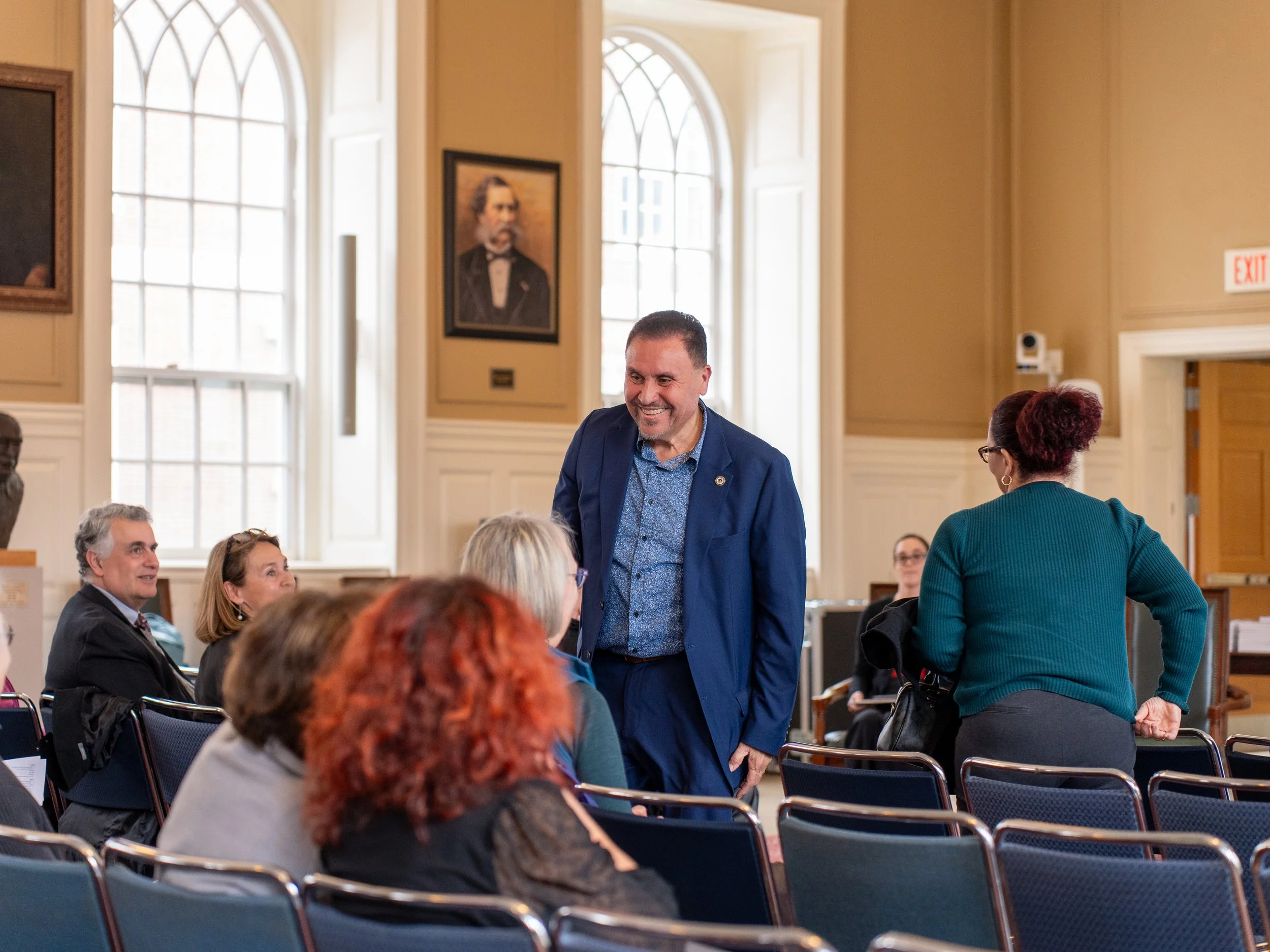A man in a blue suit smiling and talking with a woman with her back to the camera, in a large room with high windows and framed portraits on the wall. Several seated and standing people are visible.