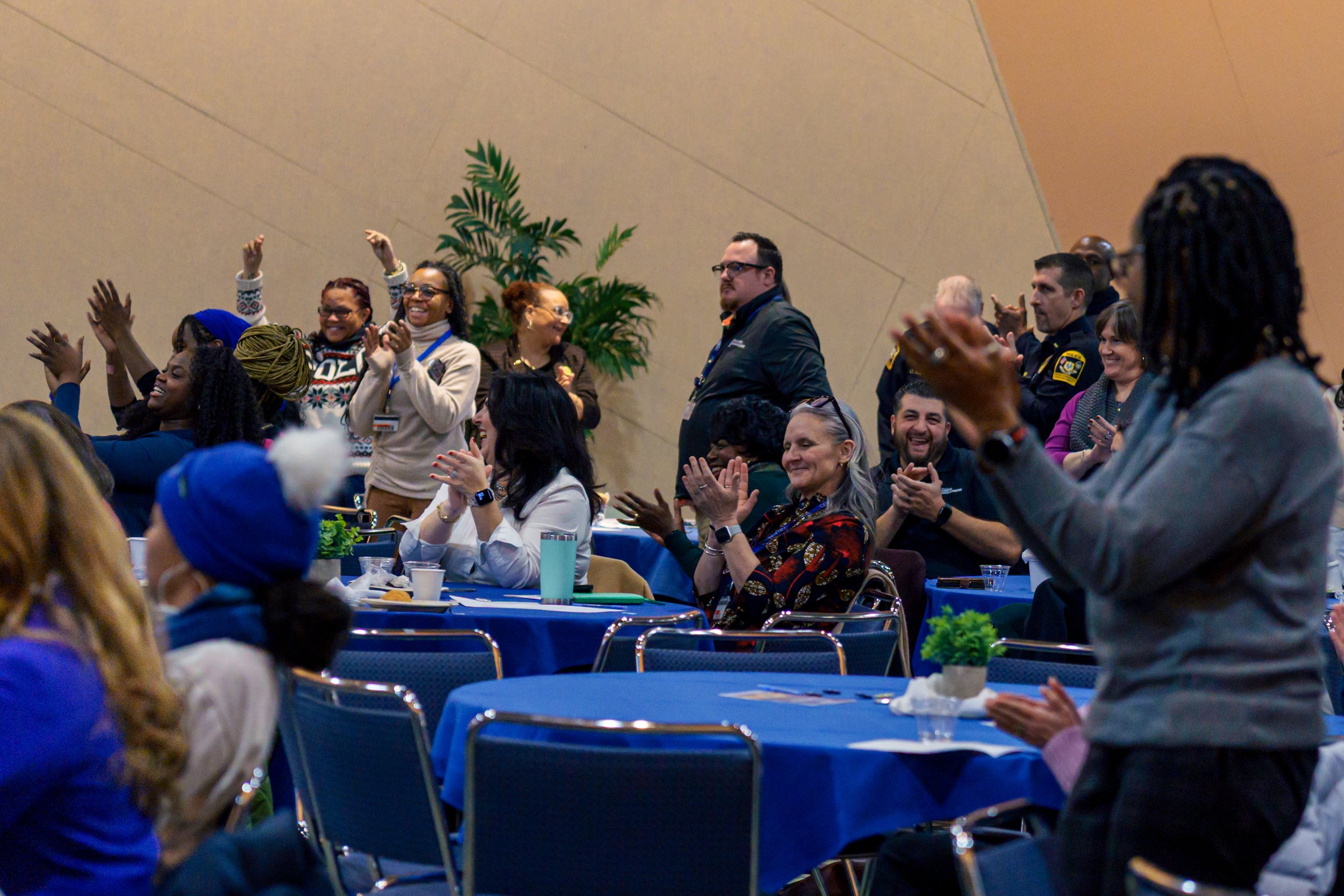 People standing and clapping at conference or event, smiling and engaging in the audience, with tables and chairs in a large indoor space.