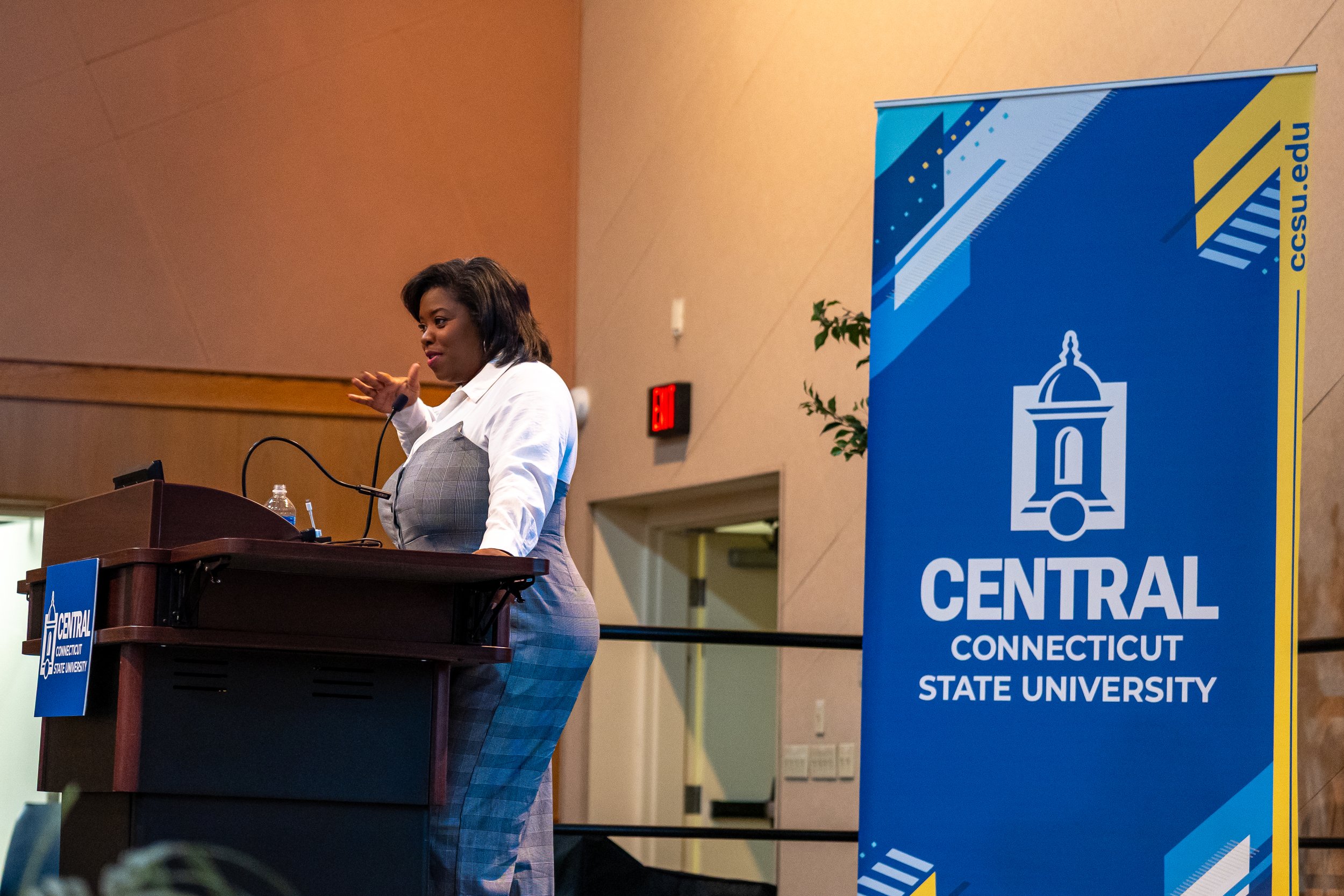 A woman giving a speech at a podium during a conference at Central Connecticut State University, with a blue and yellow CSU banner in the background.