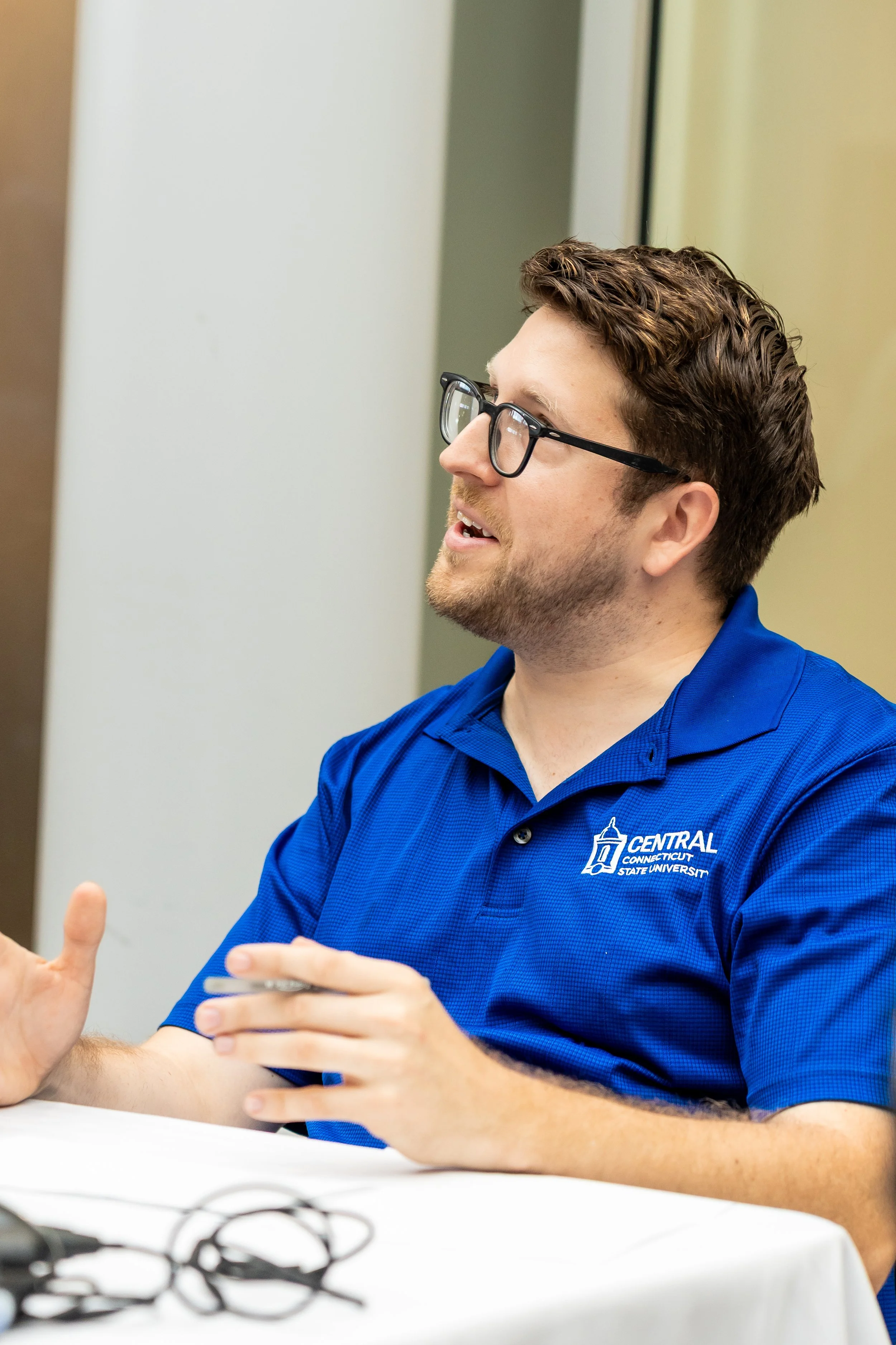 A man wearing glasses and a blue polo shirt with the logo of Central Connecticut State University is engaged in conversation at a table.