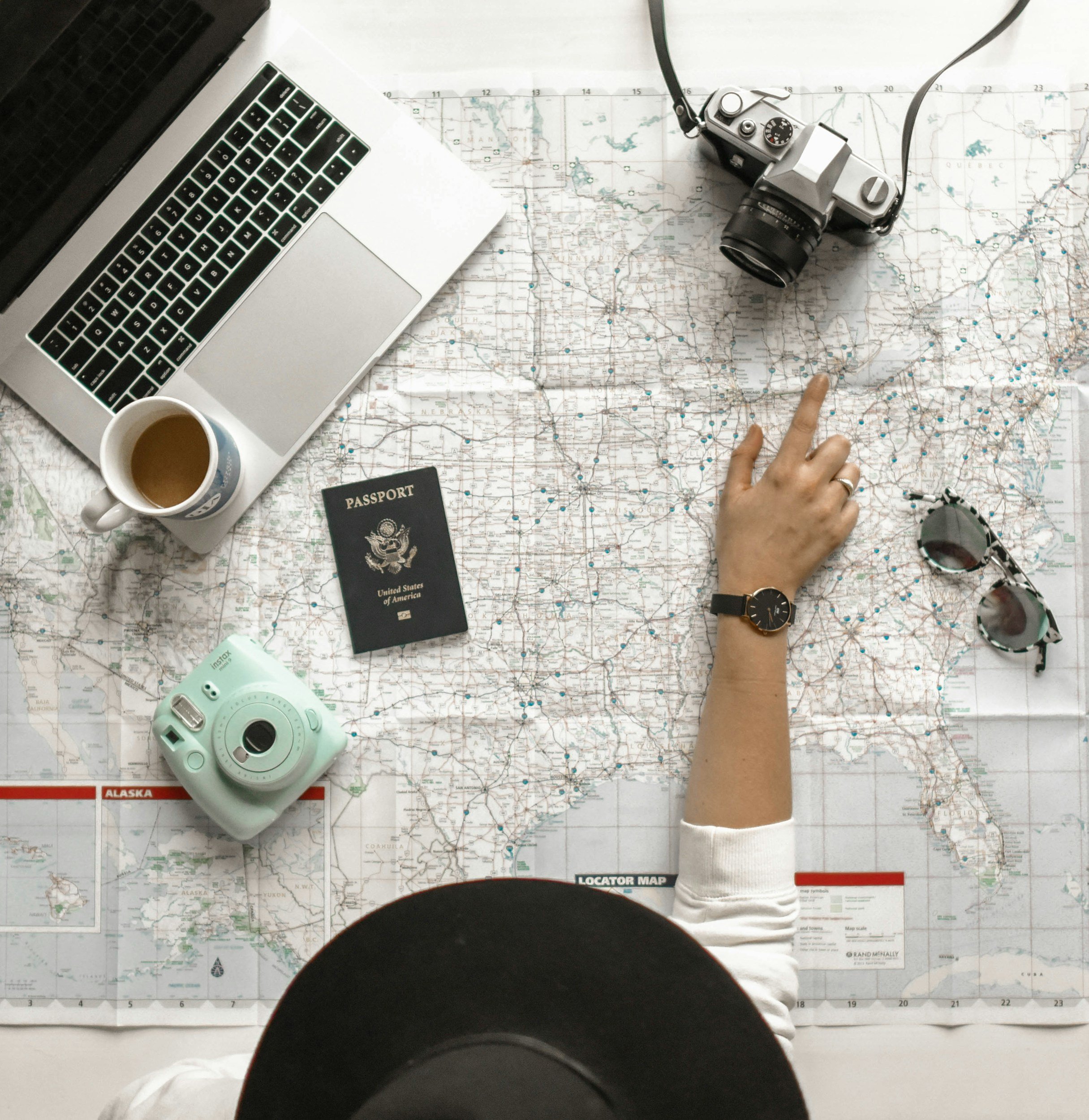 A top-down view of a person standing on a map spread on a table. The person is wearing white pants and a watch, with one finger pointing at a location on the map. Nearby are a vintage film camera, a cup of coffee, a laptop, a passport, a small printer, and a locator map.