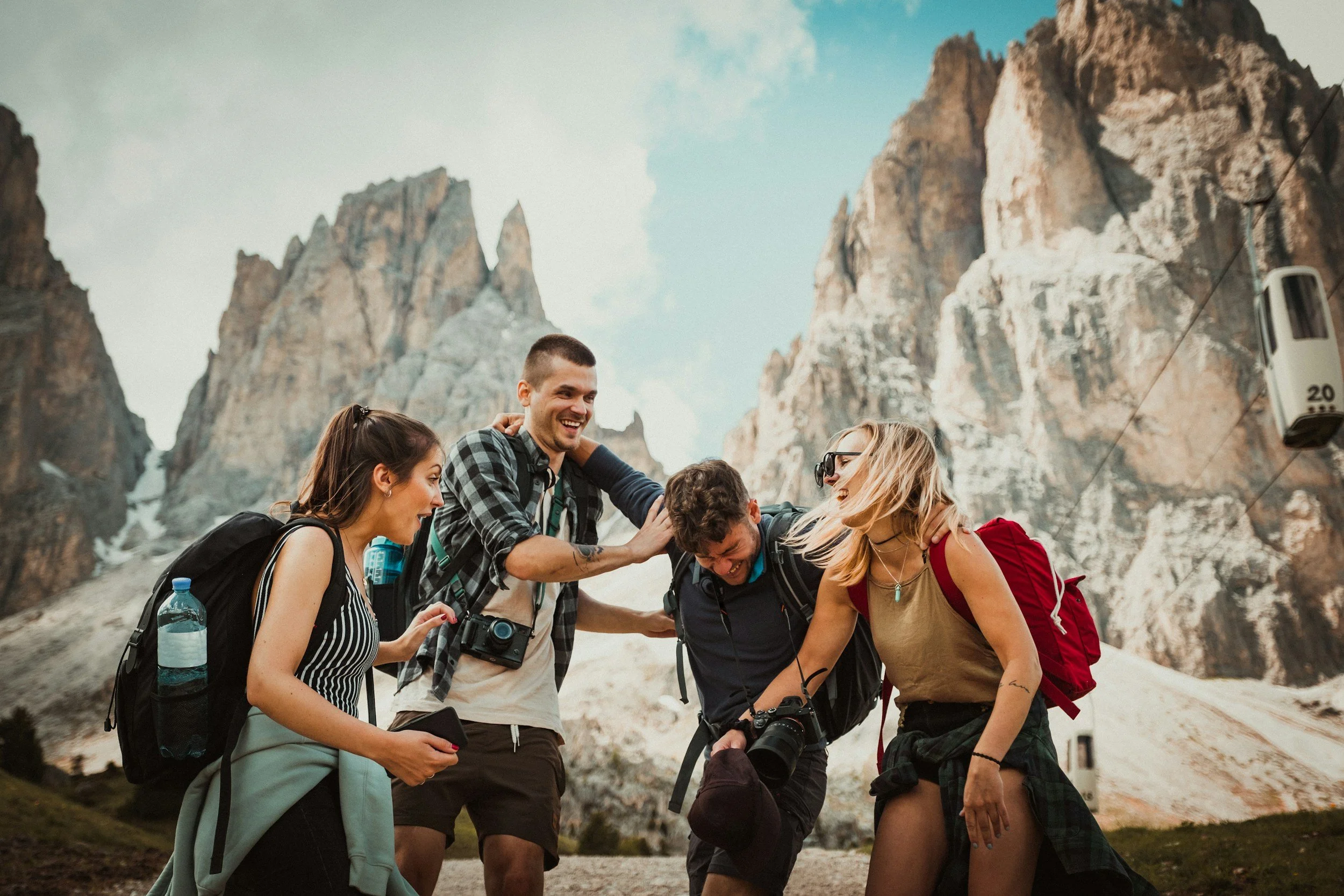 Group of five friends laughing and hiking together in a mountainous area with rocky peaks in the background, some wearing backpacks and carrying cameras.