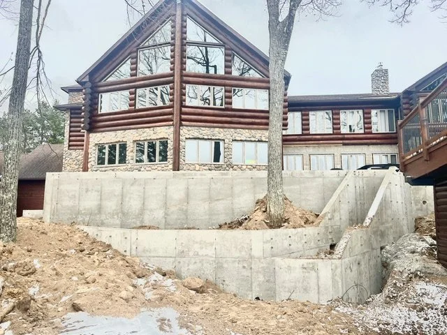 A large house with a log cabin-style exterior, multiple windows, and an expanded glass section on the upper level, built on a raised concrete foundation surrounded by trees and dirt.
