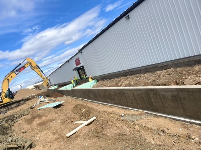 Construction site with an excavator, dirt, and an unfinished sidewalk in front of a large warehouse-looking building.
