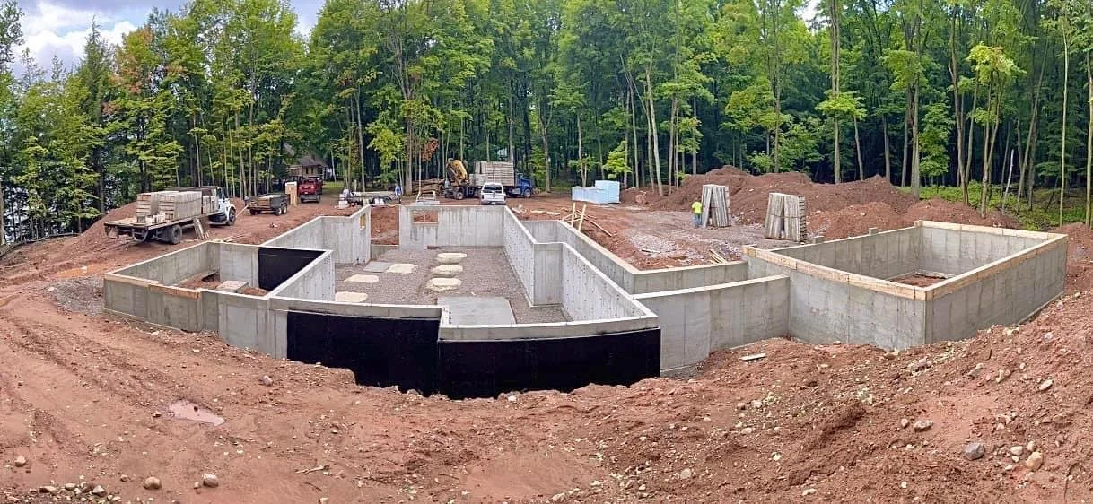 Construction site with concrete foundation in a wooded area, with vehicles and workers present.