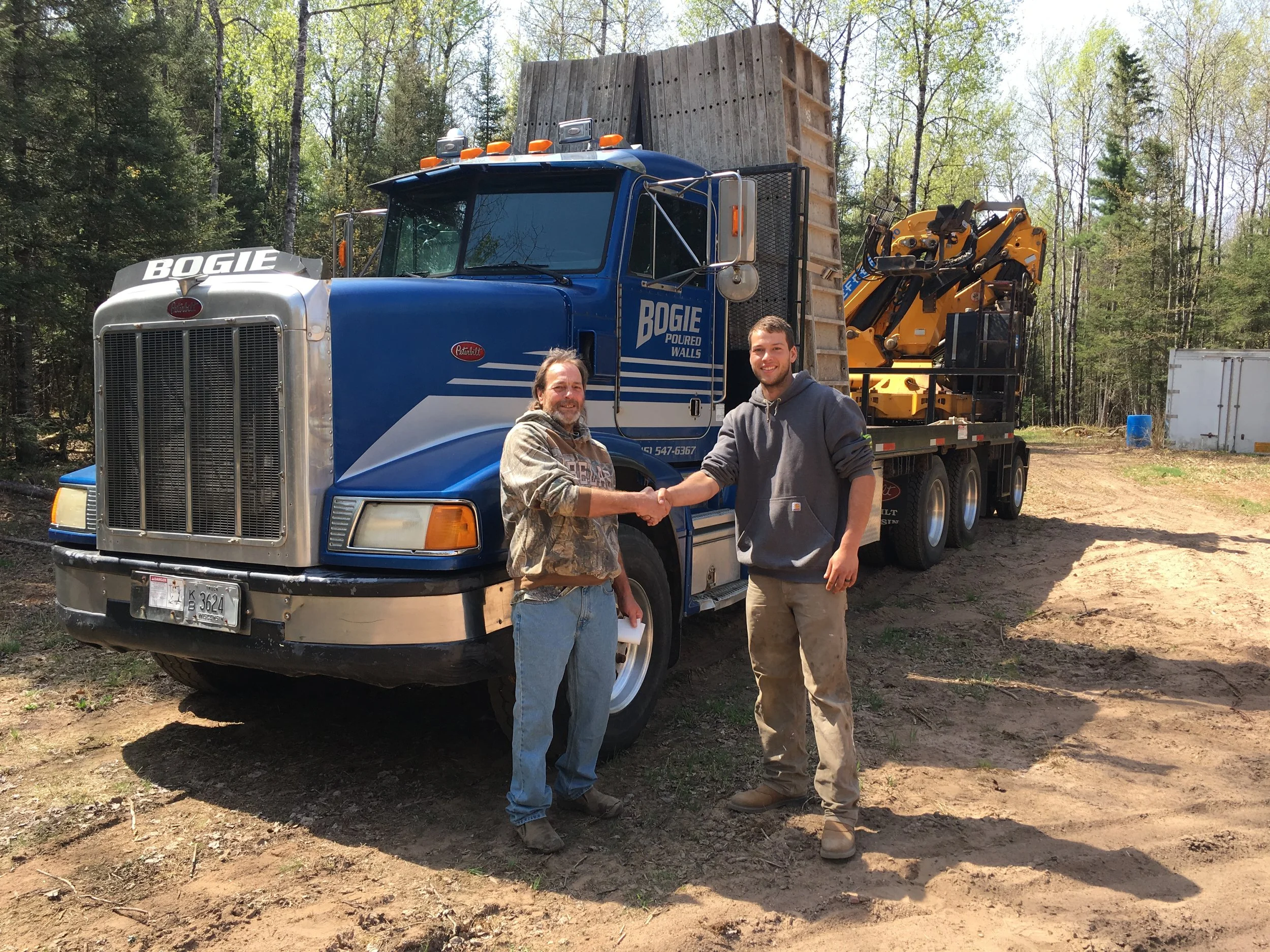 Two men shake hands in front of a large blue truck with a trench wall on its bed, in a wooded outdoor setting.