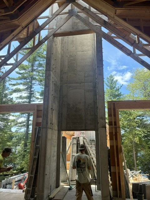 Construction site with wooden framing and concrete chimney under a roof, workers present, trees and blue sky outside.