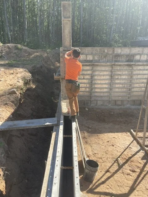 Construction worker in orange shirt and beige pants working on a large steel structure at a construction site surrounded by dirt and trees.