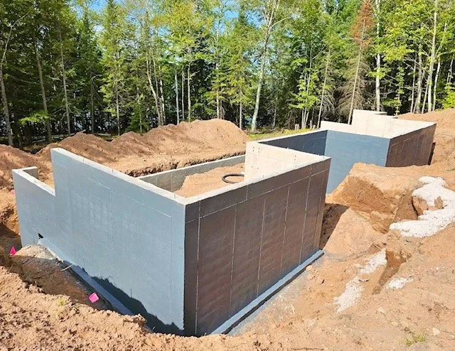 Construction site with partially built foundation walls surrounded by dirt and soil, with trees in the background.