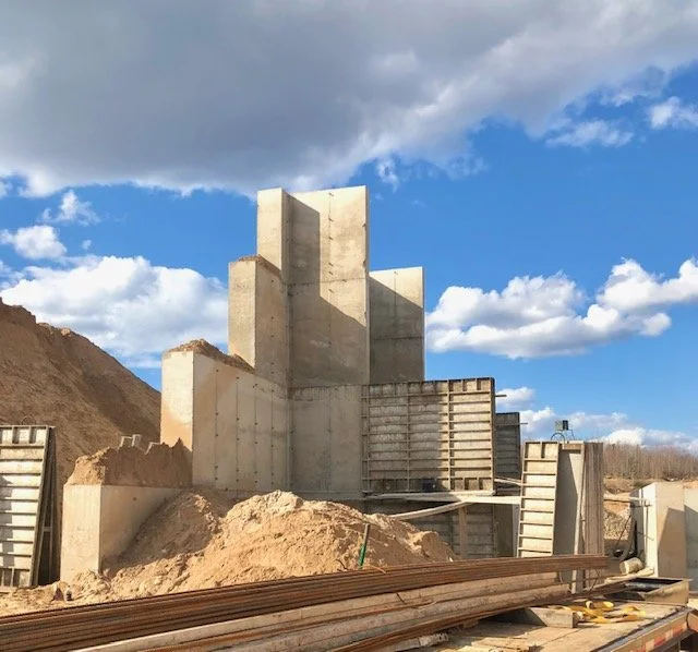 Construction site with large concrete structure under construction, wooden and metal forms, and sandy terrain, under a partly cloudy sky.