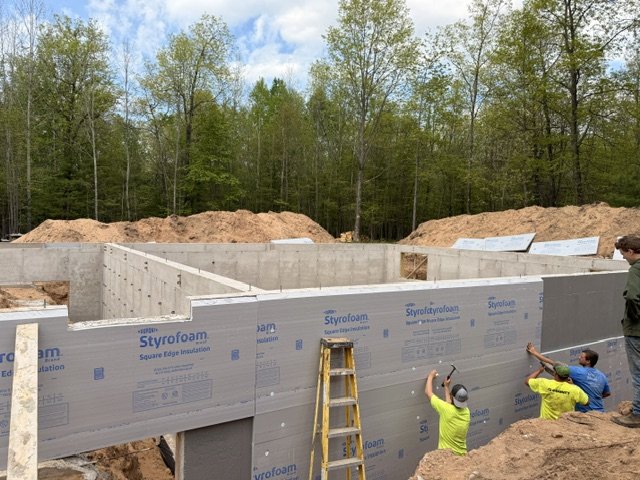 Construction workers installing Styrofoam insulation on concrete foundation walls at a building site surrounded by trees.