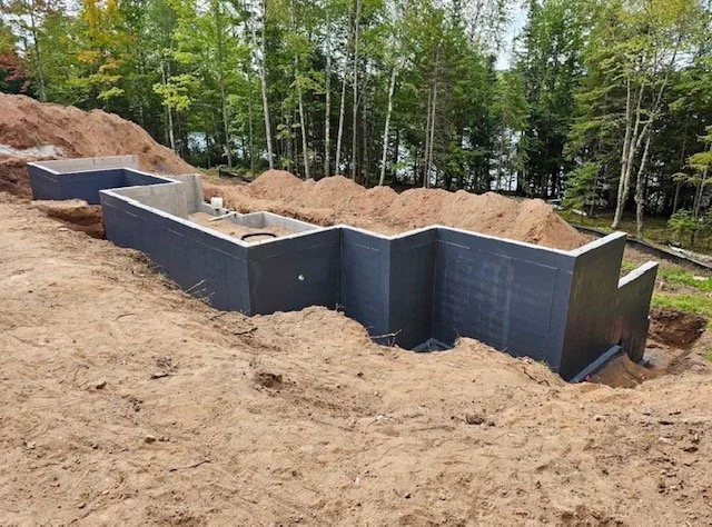 Construction site with foundation walls of a building under construction, surrounded by dirt and trees in the background.