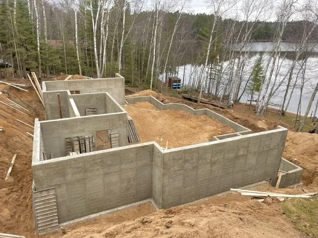Construction site on a sloped terrain with concrete foundation walls in place, surrounded by dirt, near a lake with trees in the background.