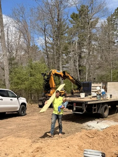A man with a child standing in front of a construction site with a flatbed truck, an excavator, and trees in the background.