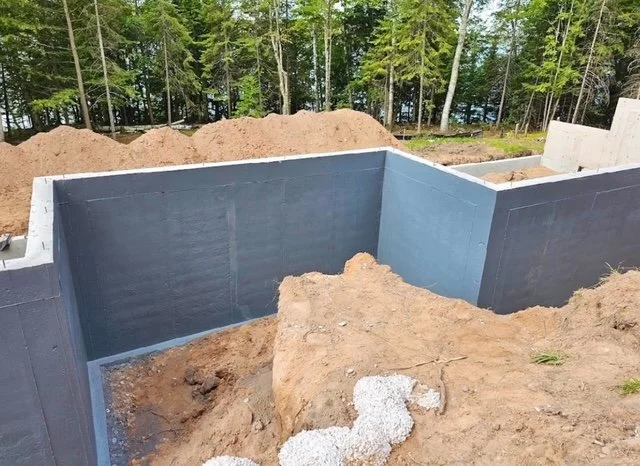 Construction site with concrete foundation walls and dirt mounds surrounded by trees.