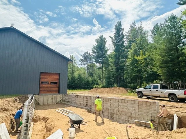 Construction site with a partially built concrete foundation, two workers, a wheelbarrow, and a pickup truck near a blue building and trees.