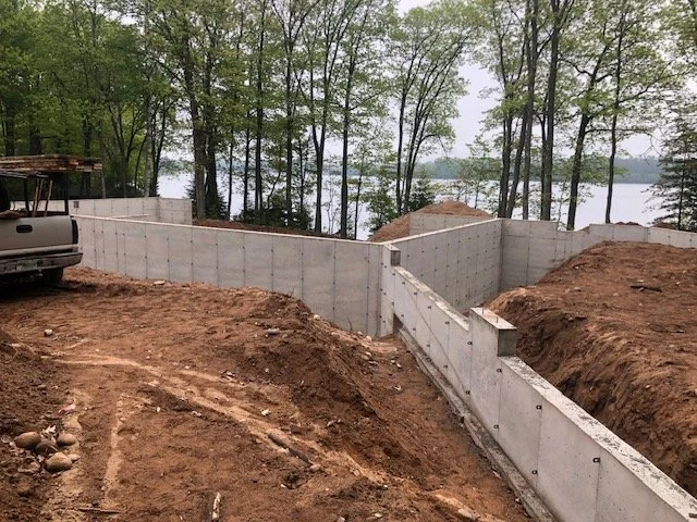 Construction site near a lake with concrete foundation walls and a parked vehicle, surrounded by trees and dirt.
