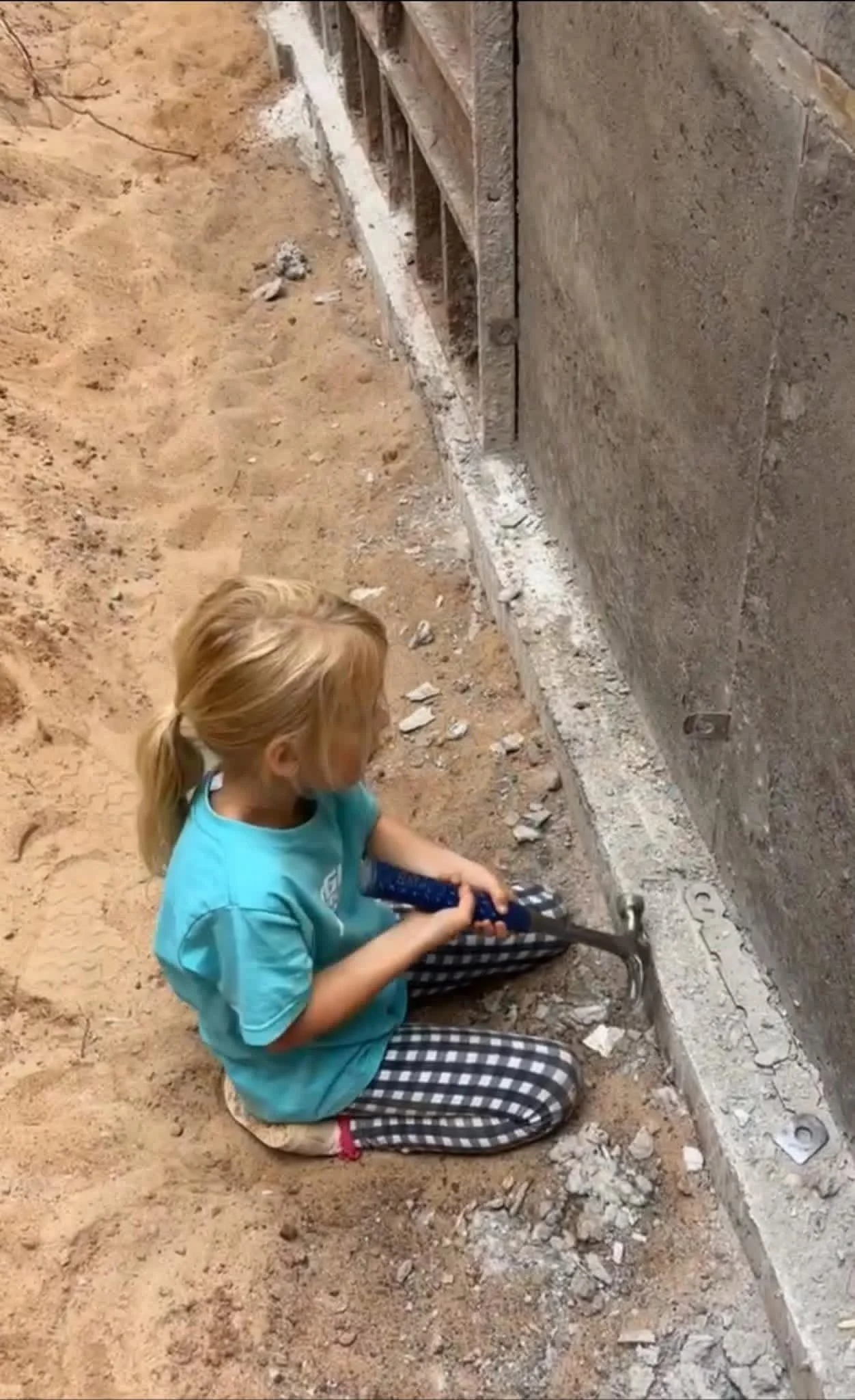 A young girl with blonde hair, wearing a blue t-shirt and checkered pants, is sitting on the ground and using a power tool to work on a concrete foundation at a construction site. The site has dirt and debris, and the girl appears to be engaged in a construction or renovation task.