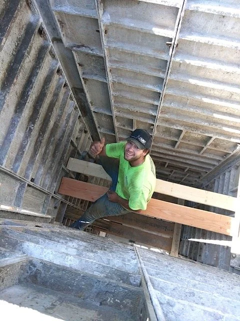 A man wearing a bright green shirt and a black cap giving a thumbs-up inside a construction site with metal and wooden framing.