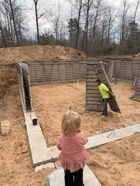 Construction site with concrete foundation and form walls in progress, two children observing, one boy holding a form panel, trees in background.