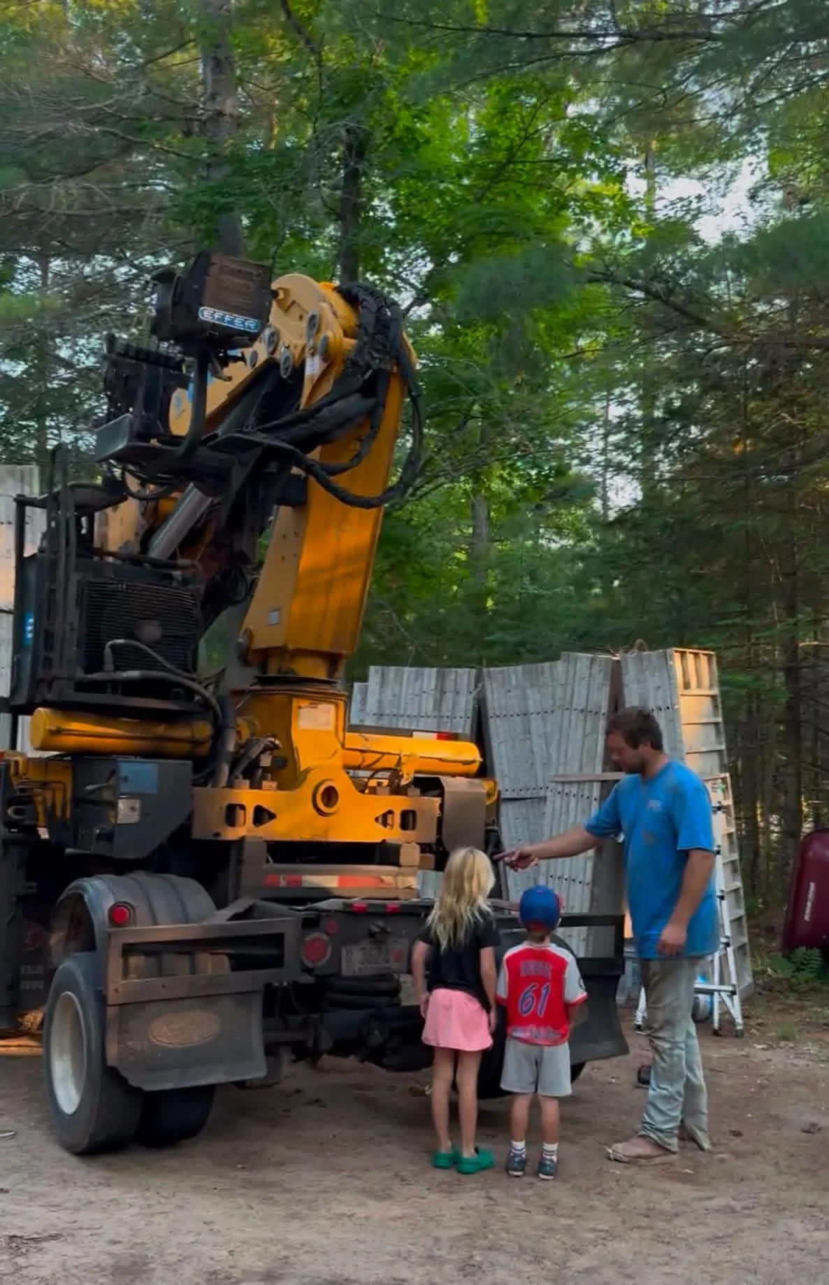 A large yellow construction machine with a hydraulic arm is parked on a dirt surface, surrounded by trees. Two children and an adult are standing in front of the truck, interacting with a person who appears to be a worker.