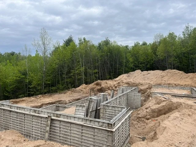 Construction site with wooden formwork for a foundation, surrounded by excavated dirt and a forested area in the background.