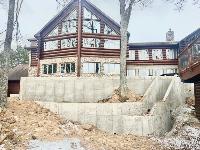 A large house built on a sloped terrain with a stone foundation, wooden log exterior, and large windows, surrounded by trees, with a concrete retaining wall and steps leading down to the yard.