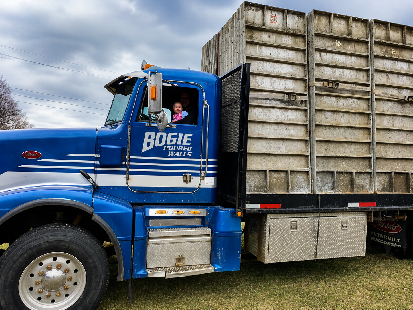 A large blue Peterbilt dump truck with two children in the driver’s seat, smiling. The truck is labeled "Rogie Poured Walls" on the side, parked outdoors on a grassy area with cloudy skies in the background.
