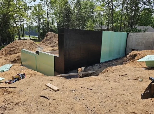 Construction site with partially built concrete foundation wall, surrounded by dirt and construction materials.