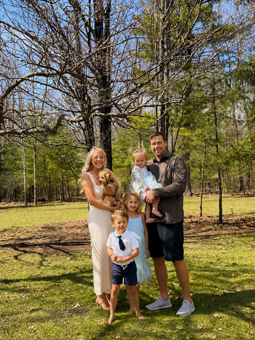 Family of five with a dog posing outdoors in a grassy area with trees and a blue sky.