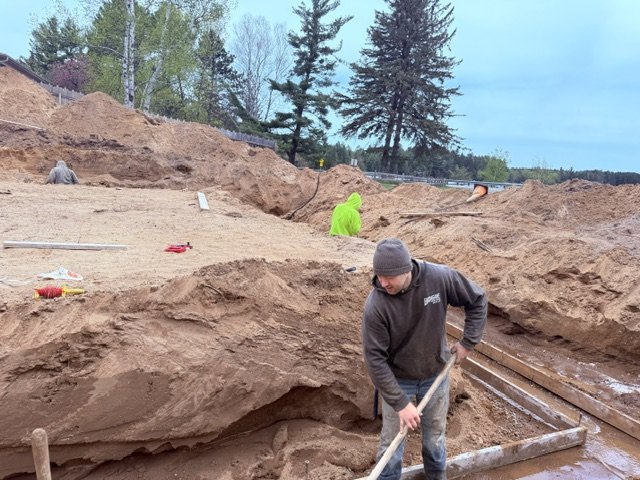 Three men working at an archaeological excavation site with dirt, tools, and trenches, surrounded by trees and a blue sky.