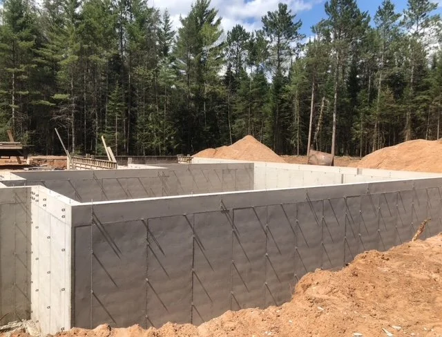 Construction site showing concrete foundation with black waterproofing membrane and steel reinforcement grid, surrounded by dirt and trees in the background.
