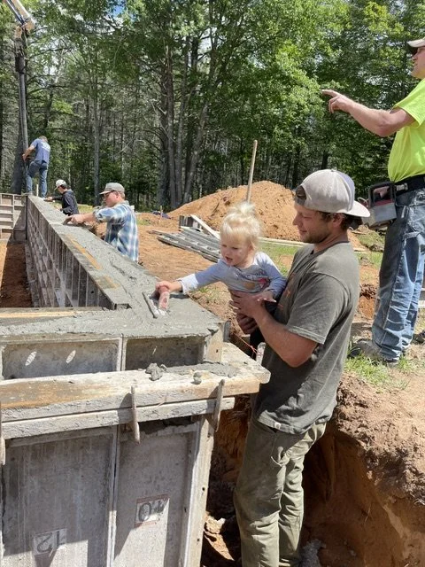A construction site with several people, including a man holding a young girl, working on a concrete structure. The scene is outdoors, surrounded by trees and dirt piles.