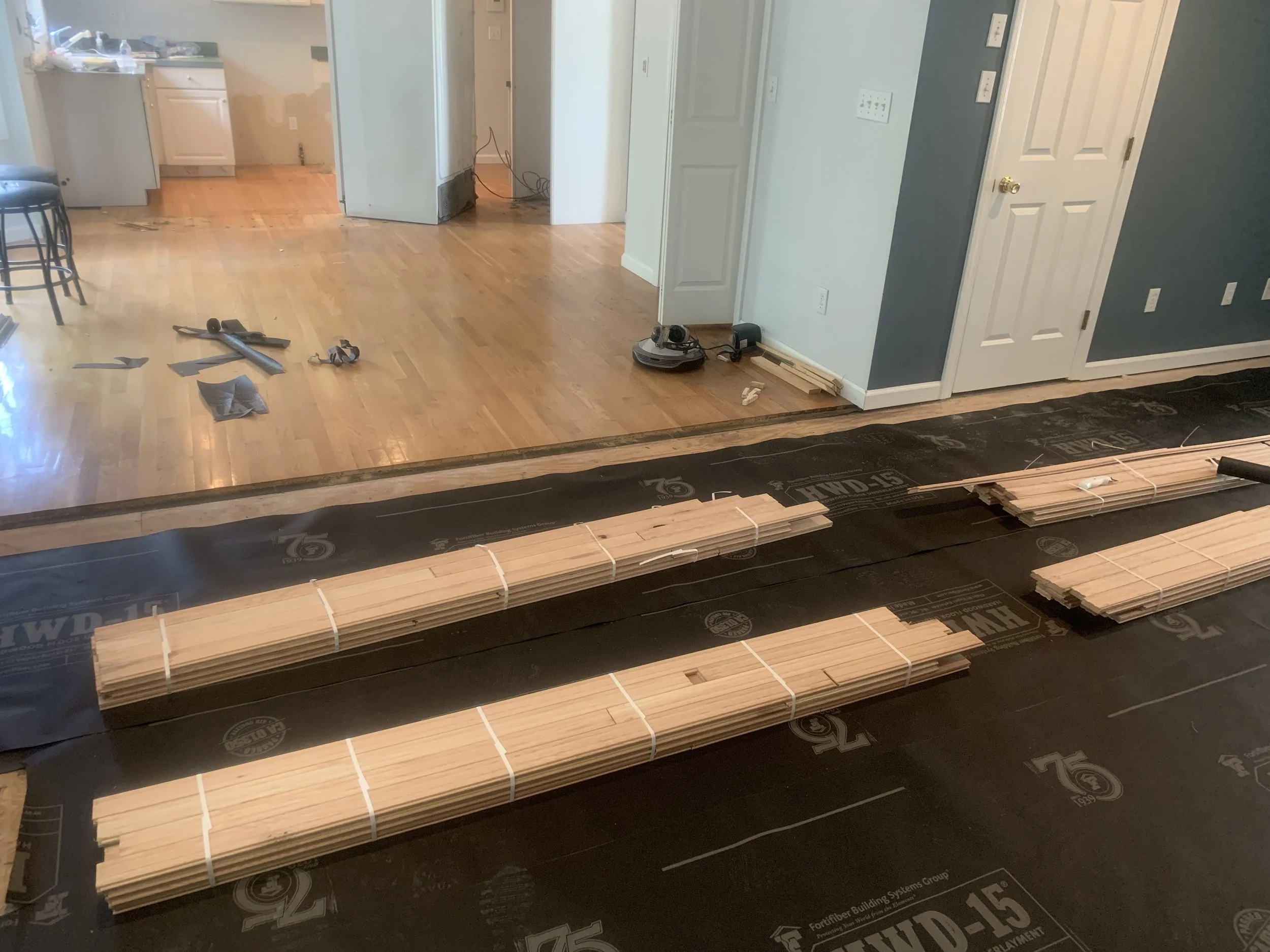 Living room with hardwood floor during renovation, with tools and flooring materials scattered on the floor, and some walls painted blue and white.
