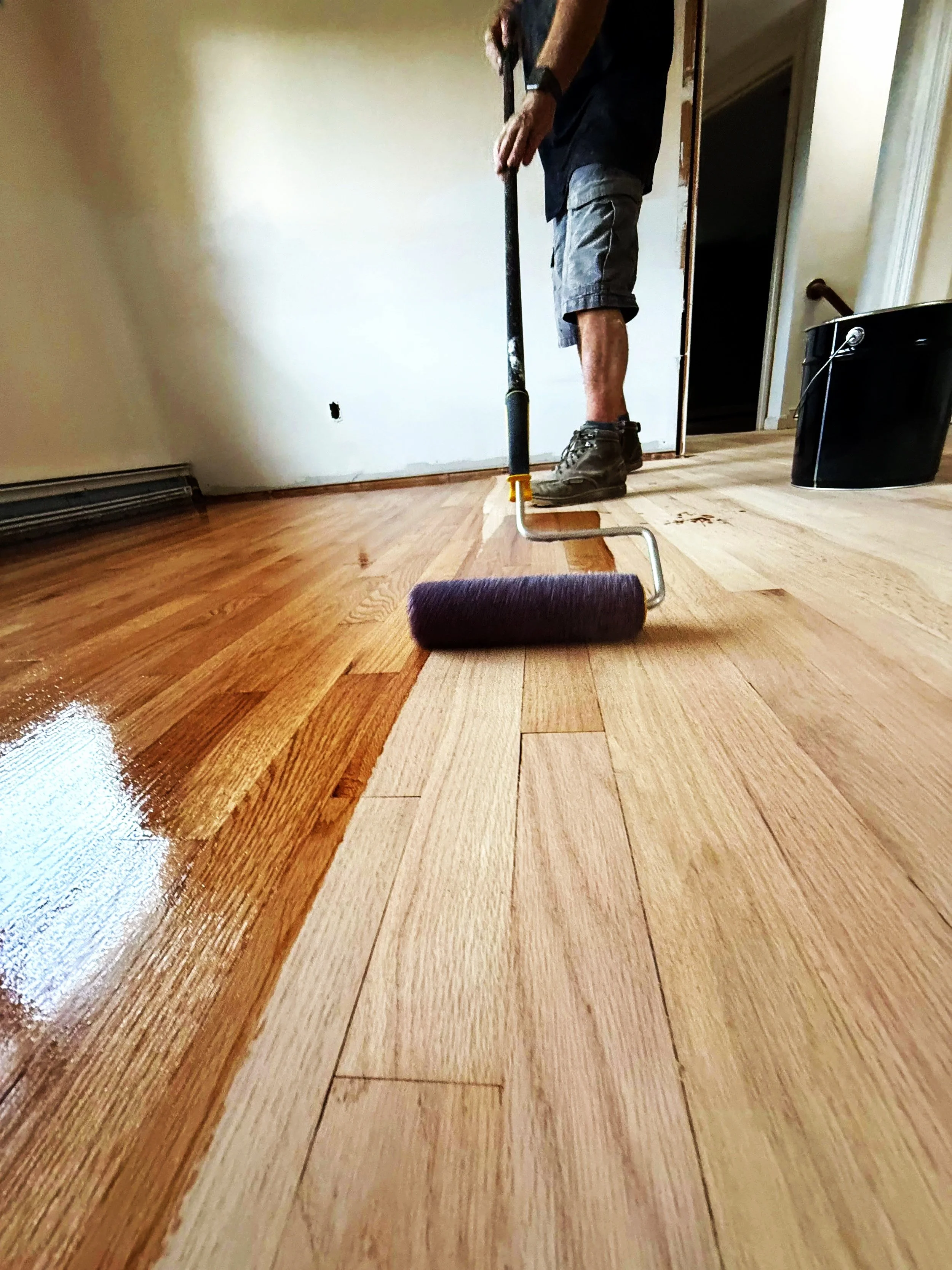 Person applying stain or finish to a wooden floor with a roller brush.