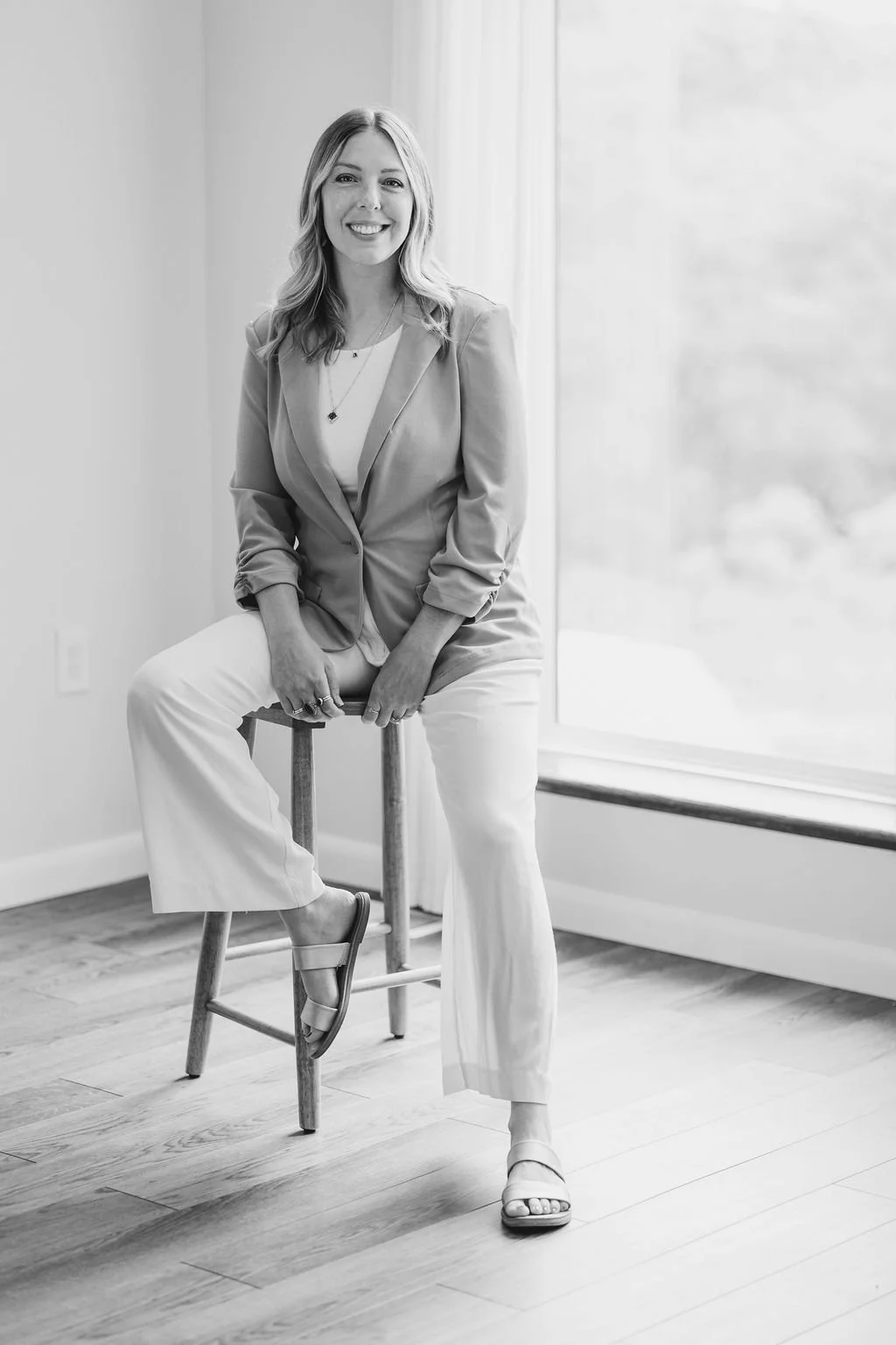 A woman with wavy shoulder-length hair smiling, sitting on a stool near a large window, wearing a blazer, white shirt, white pants, and sandals in a bright room.