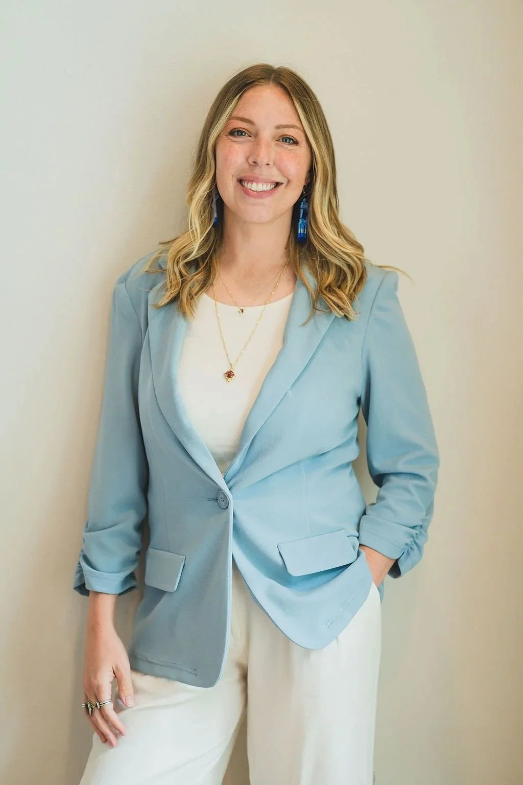 A woman with blonde, wavy hair smiling, wearing a light blue blazer over a white top, with blue earrings, layered necklaces, and beige pants, standing against a plain wall.