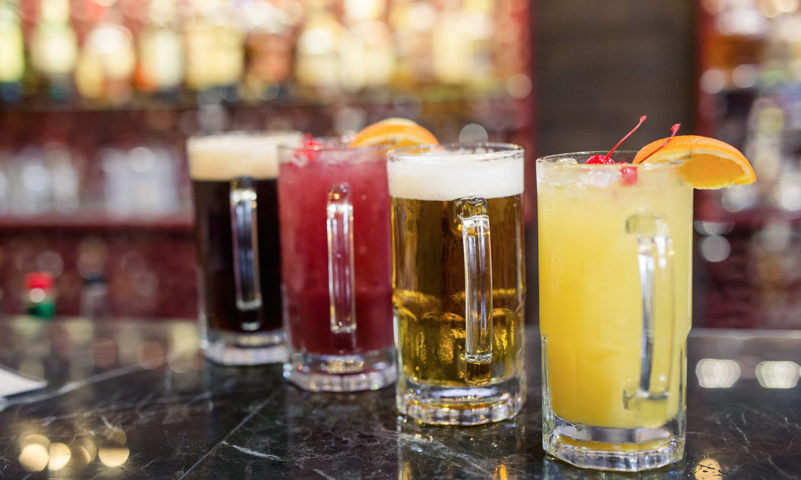 Four colorful cocktails on a bar counter, garnished with fruit slices and cherries, with a blurred bar background.