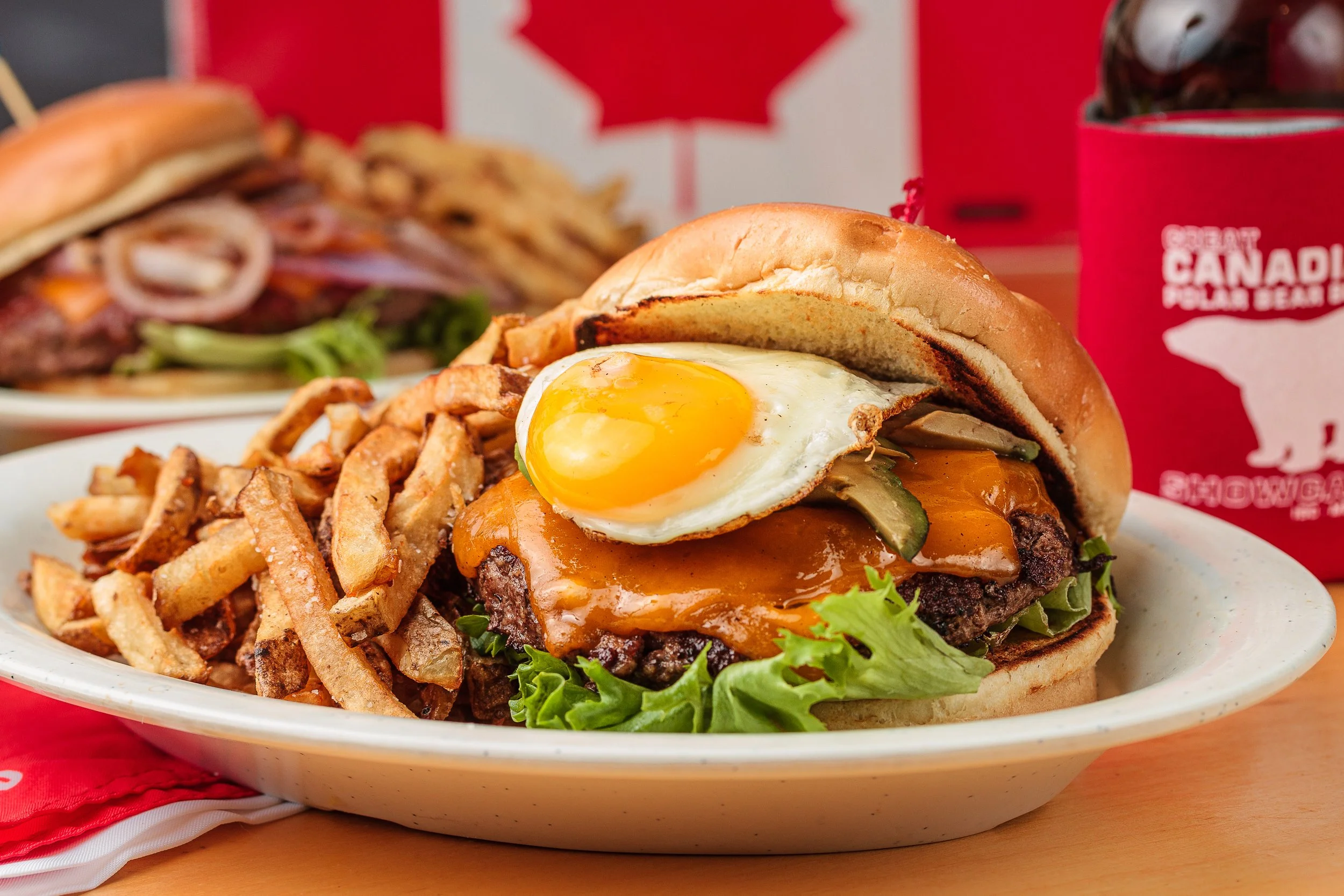 A plate with a burger containing lettuce, a fried egg, cheese, and a beef patty, served with French fries. In the background, there are Canadian-themed items and a Canadian flag.