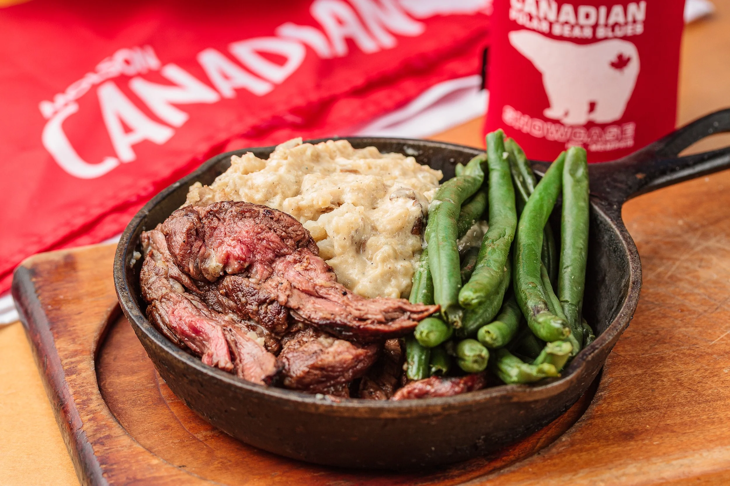 A cast iron skillet containing sliced medium-rare roast beef, creamy mashed potatoes, and green beans, placed on a wooden tray with a red Canadian Bear Logo in the background.