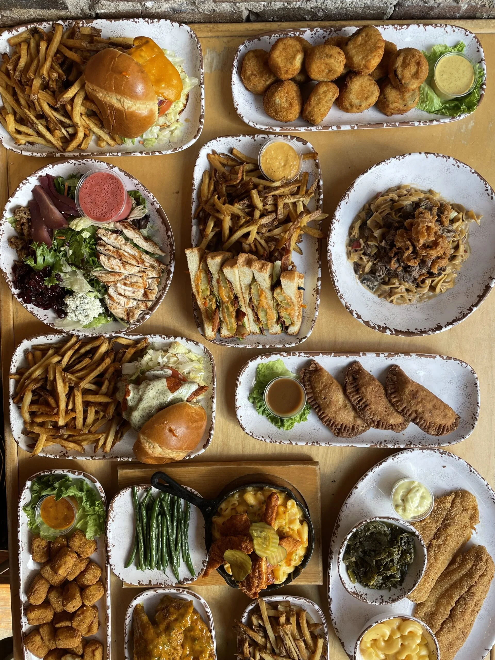 A variety of dishes served on trays, including French fries, fried chicken, various salads, sandwiches, egg rolls, and other entrees, with some dipping sauces and condiments.