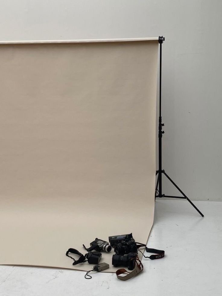 Photography backdrop stand with a beige backdrop and camera equipment, including cameras and straps, placed on the floor.