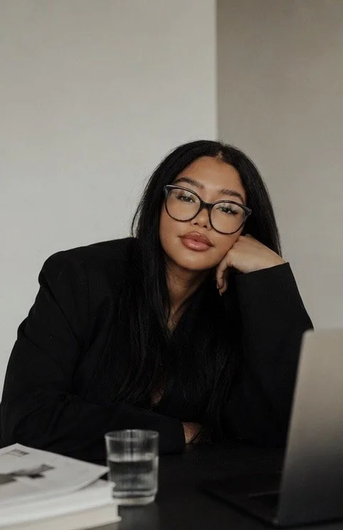 A woman with long black hair, glasses, and a black blazer sitting at a table with a laptop, a glass of water, and some papers, resting her head on her hand.