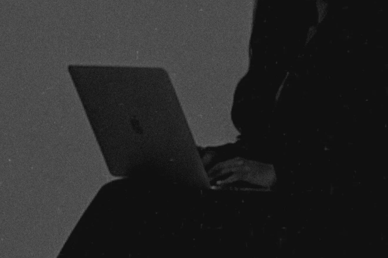 A person sitting and using a laptop in a dark environment, with the screen illuminating their hands.
