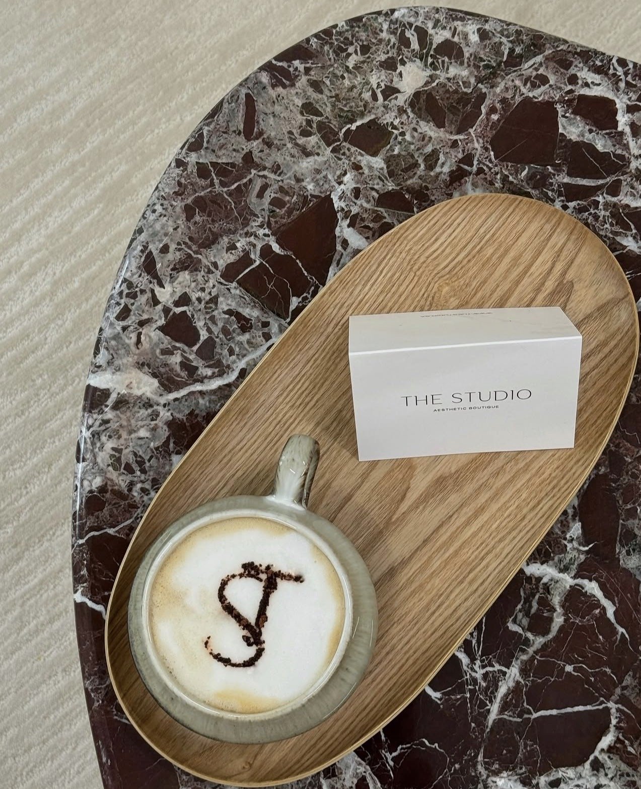 A marble table with a wooden tray holding a cup of coffee with a heart and abstract design made with cocoa powder, a business card from 'The Studio' aesthetic boutique, and a white card with black text.
